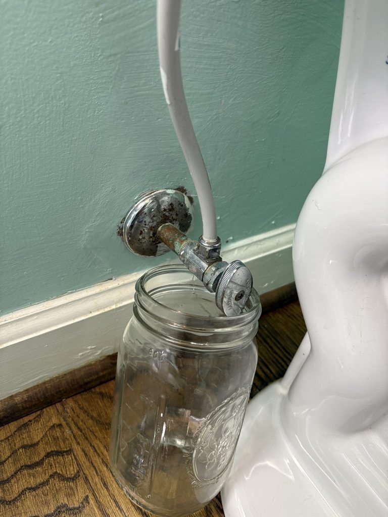 BEFORE — the toilet's shutoff, leaking into a jar | Leaking toilet shutoff valve dripping into a jar in an Apex, NC guest bathroom